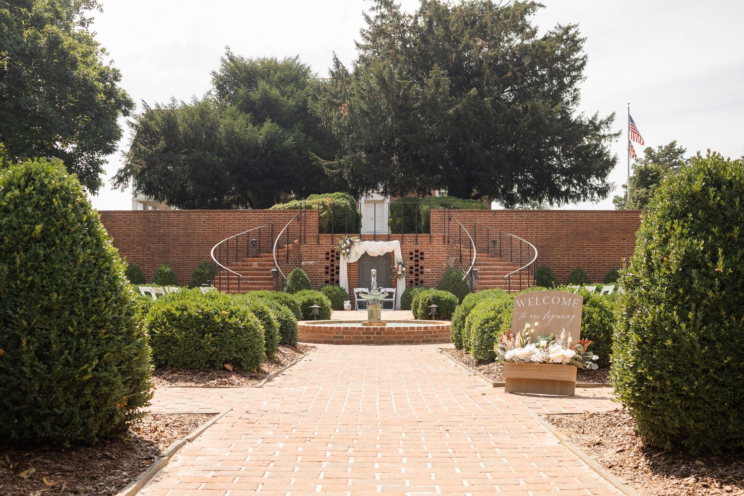 A panoramic view of a formal garden pathway with trimmed hedges and ornamental features leading to a brick structure.