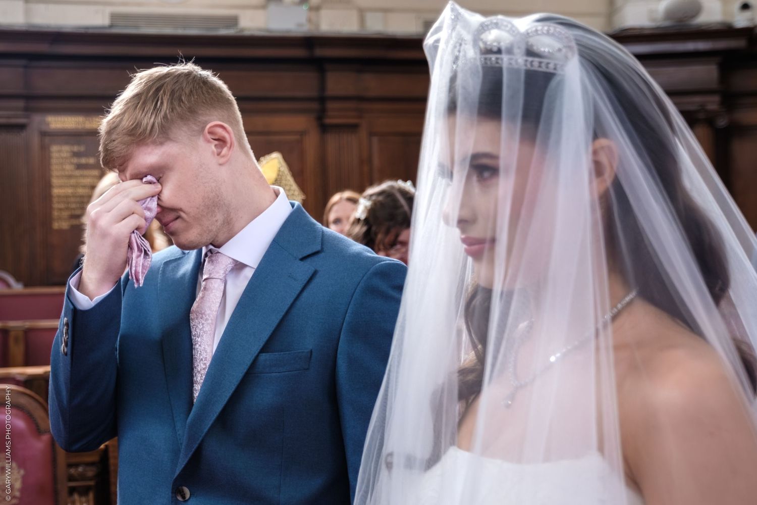 A tender moment between a couple in wedding attire, with white veil and navy suit, during a church ceremony.
