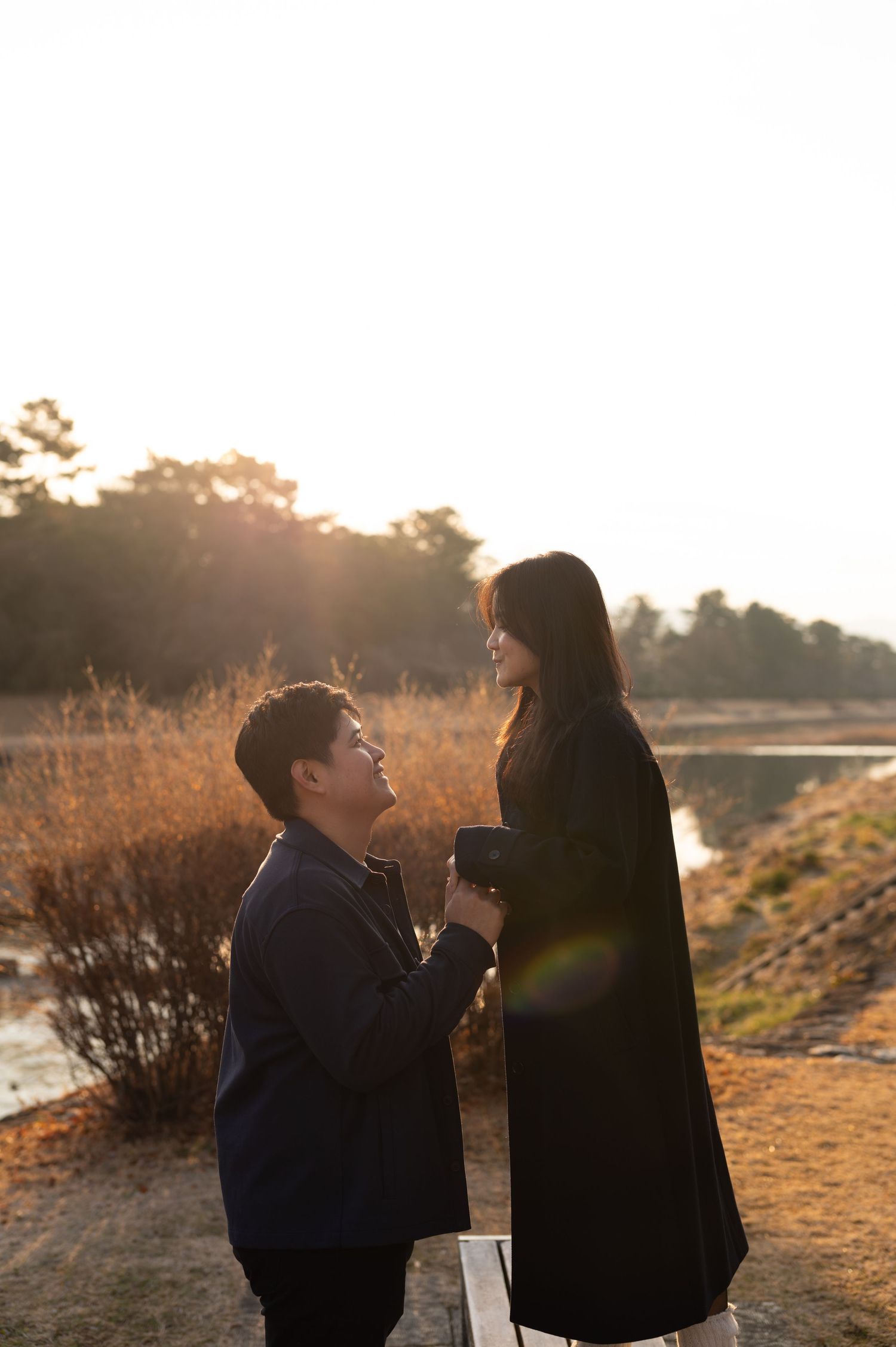 Man proposing on one knee to woman at golden sunset near river with warm lens flare.