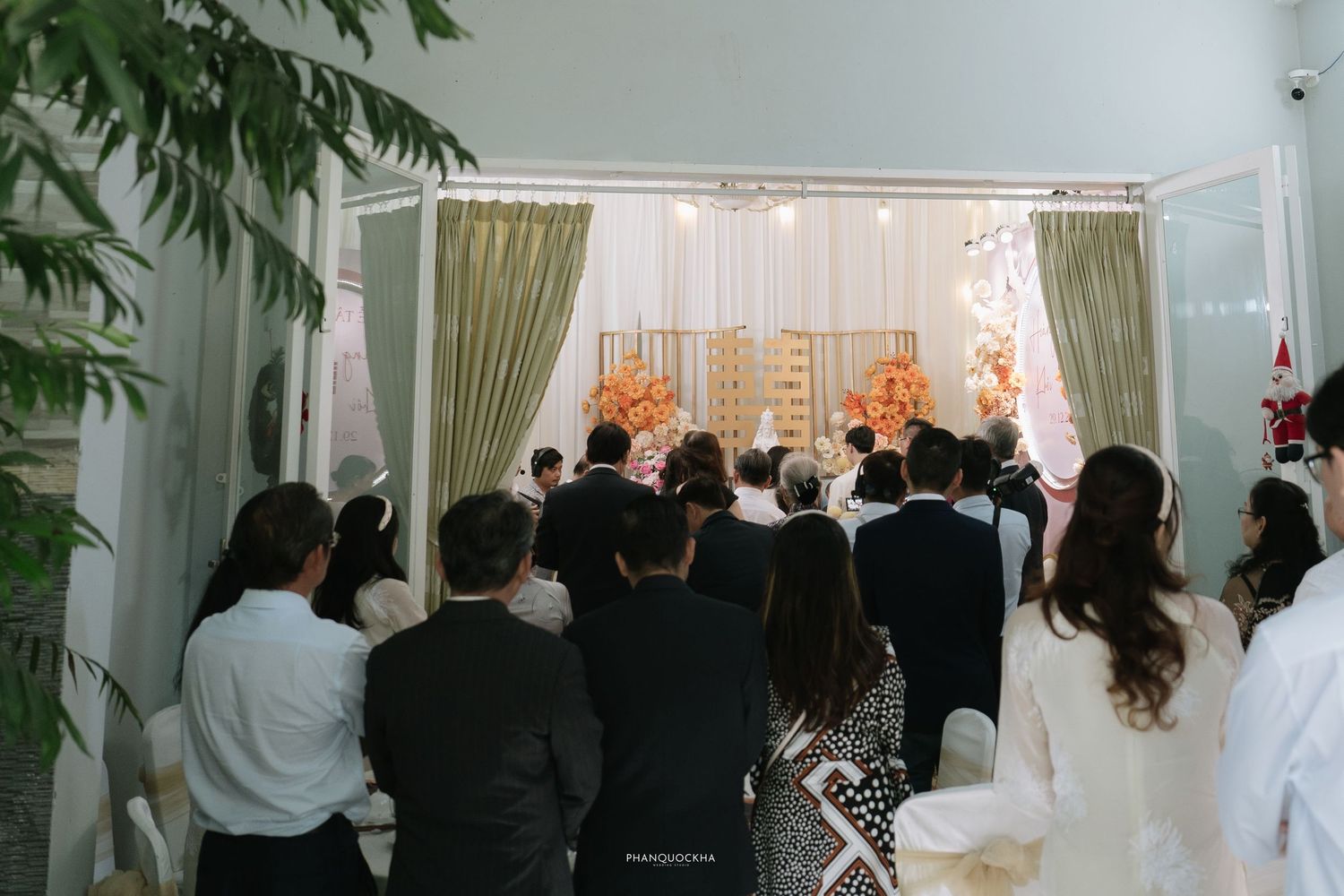 Guests stand facing an elegant wedding altar with curtains and orange floral decorations.