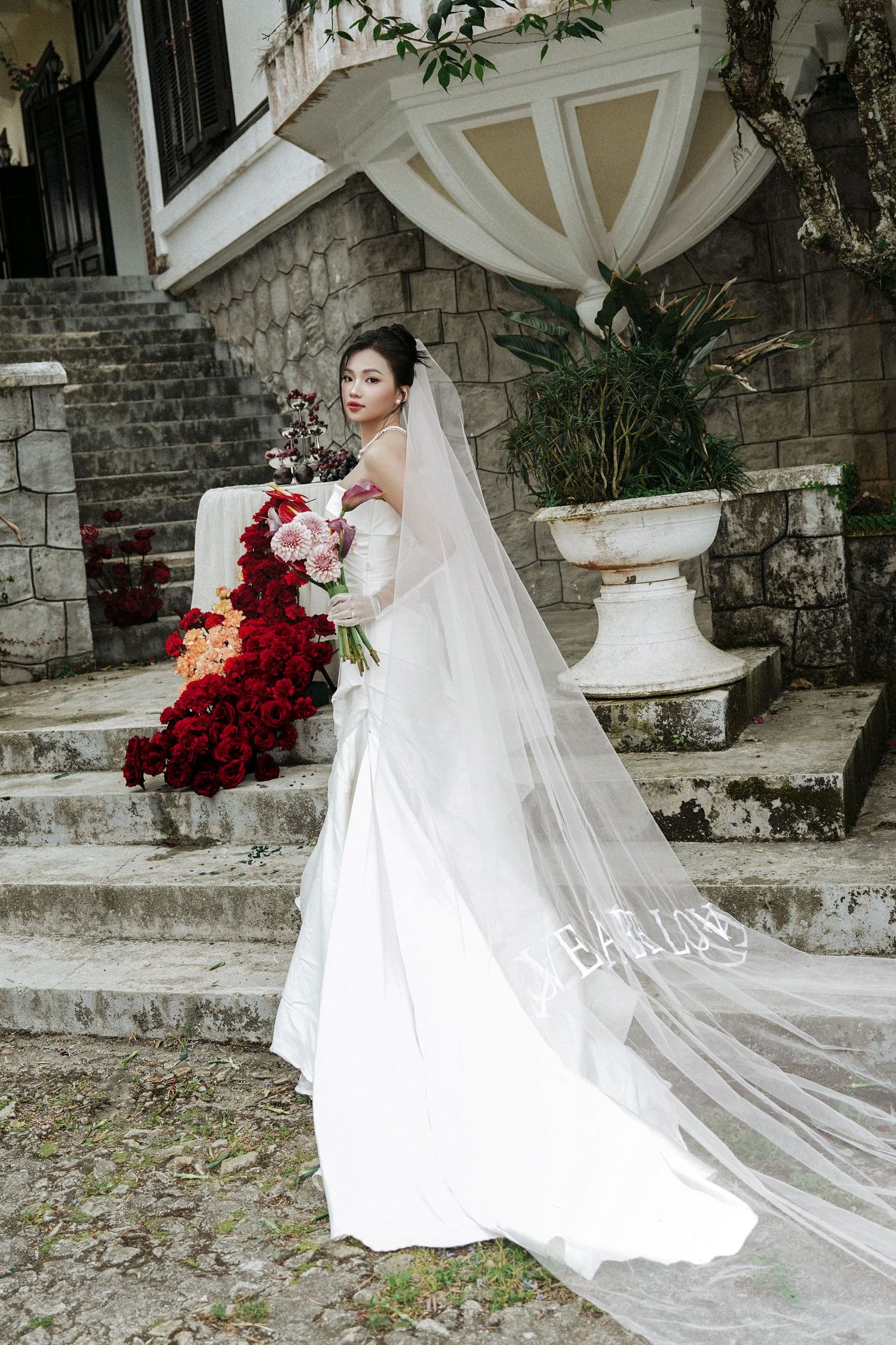 Bride in flowing white wedding gown with long veil holding bouquet on stone building staircase.