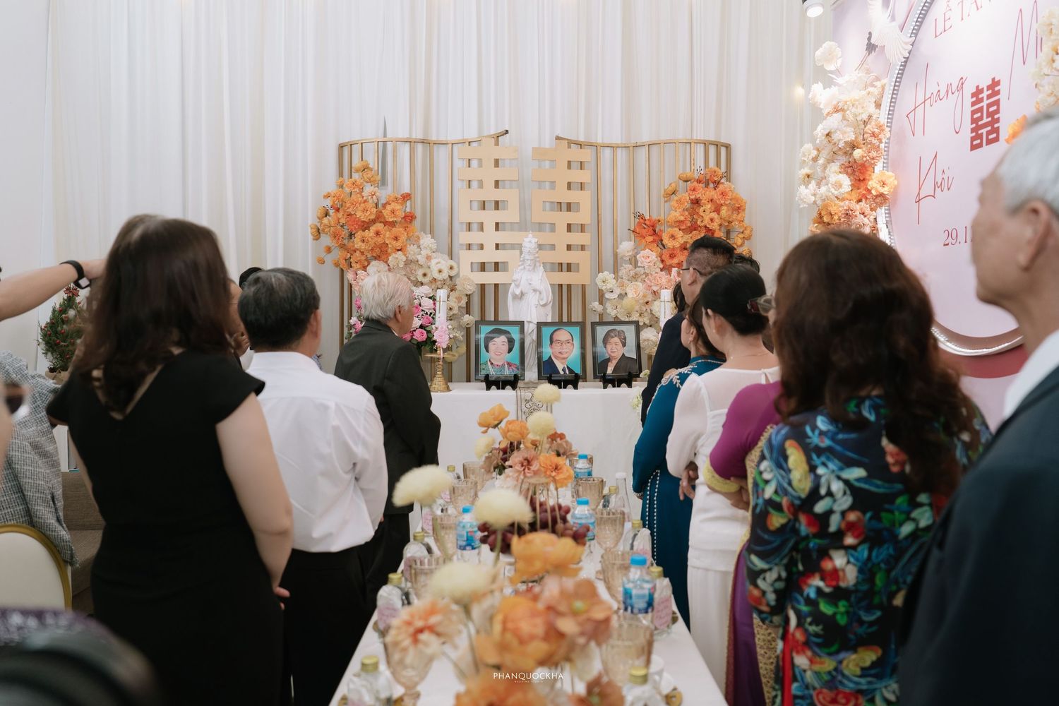 Guests gather around a decorated altar with framed photos and orange floral arrangements at a Vietnamese wedding ceremony.