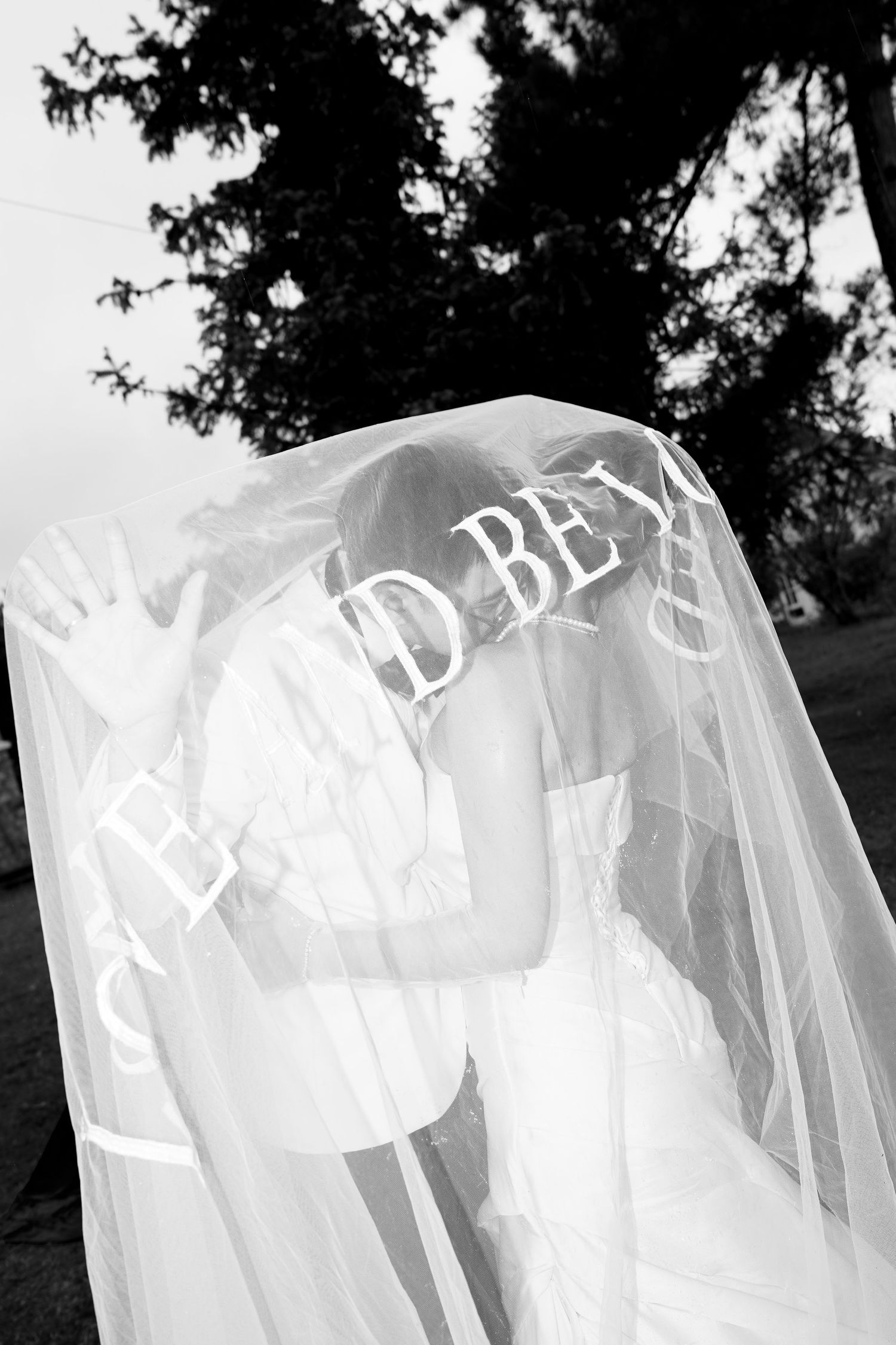 Bride and groom share a romantic kiss beneath a flowing white veil with cursive text outdoors.
