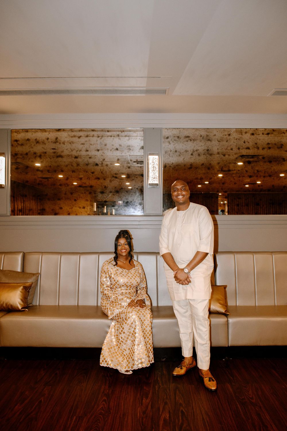 A couple poses in formal attire against a cream-colored wall with rustic wood accents.