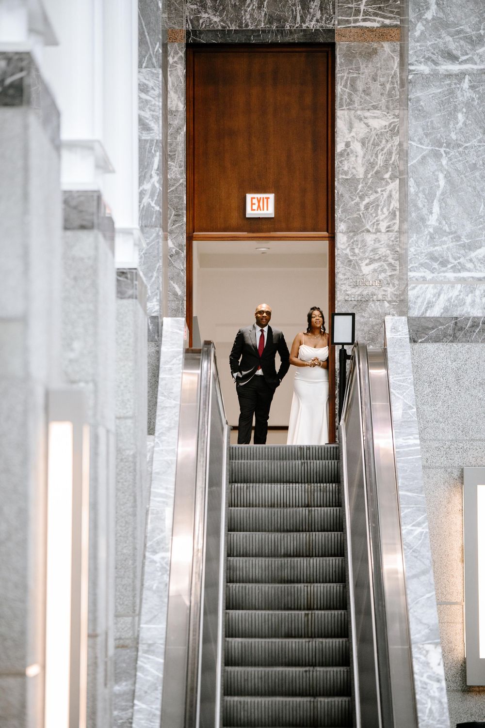 Wedding couple poses on escalators in an elegant marble-walled building with classic architectural features.