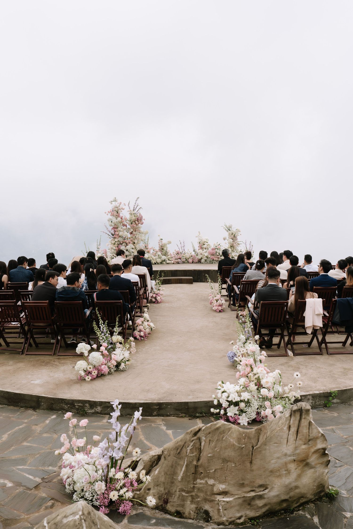 Elegant outdoor wedding ceremony on a misty hilltop with floral arrangements lining the aisle and guests seated in wooden chairs.