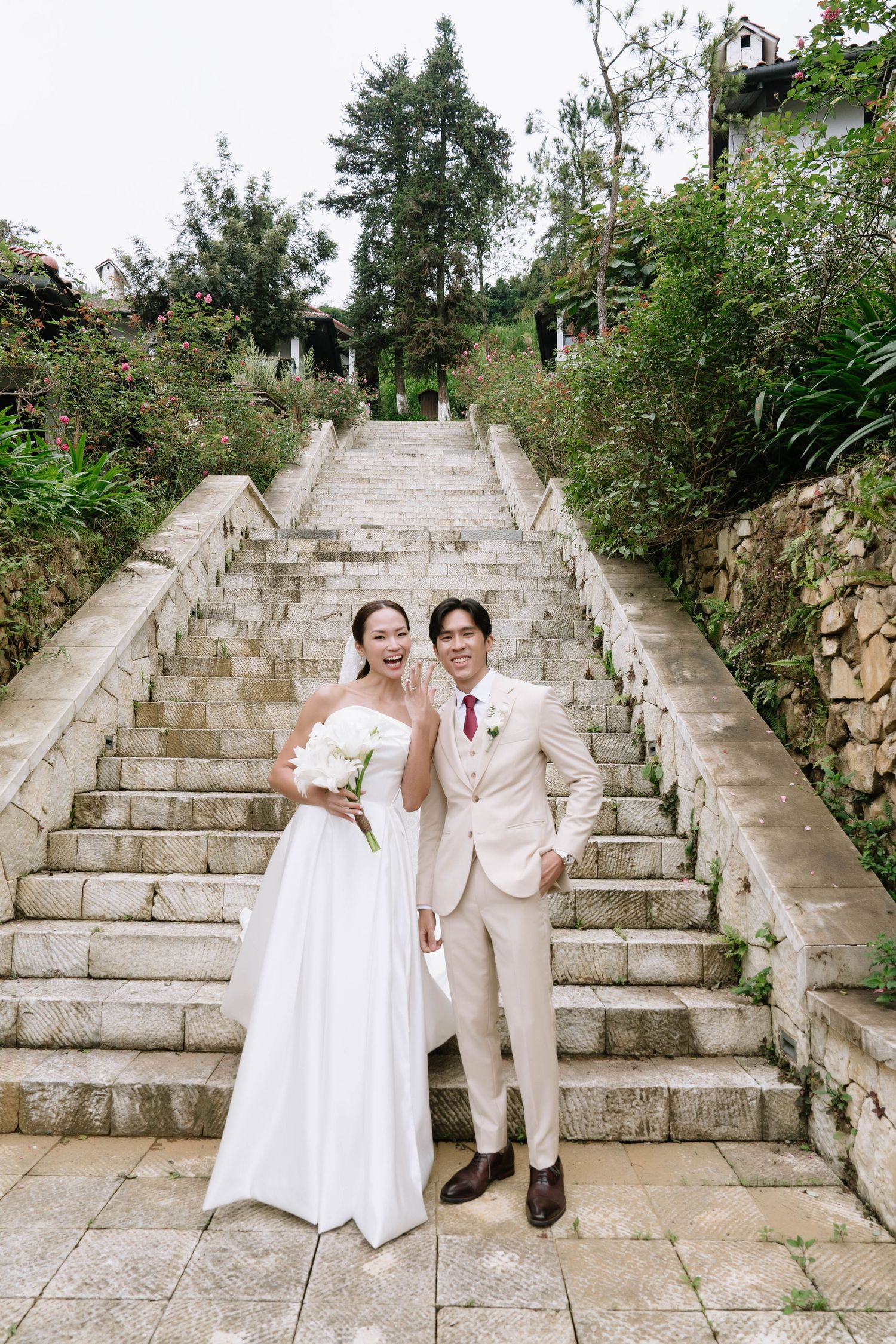 Happy Asian couple in wedding attire pose together at the base of a grand stone staircase.