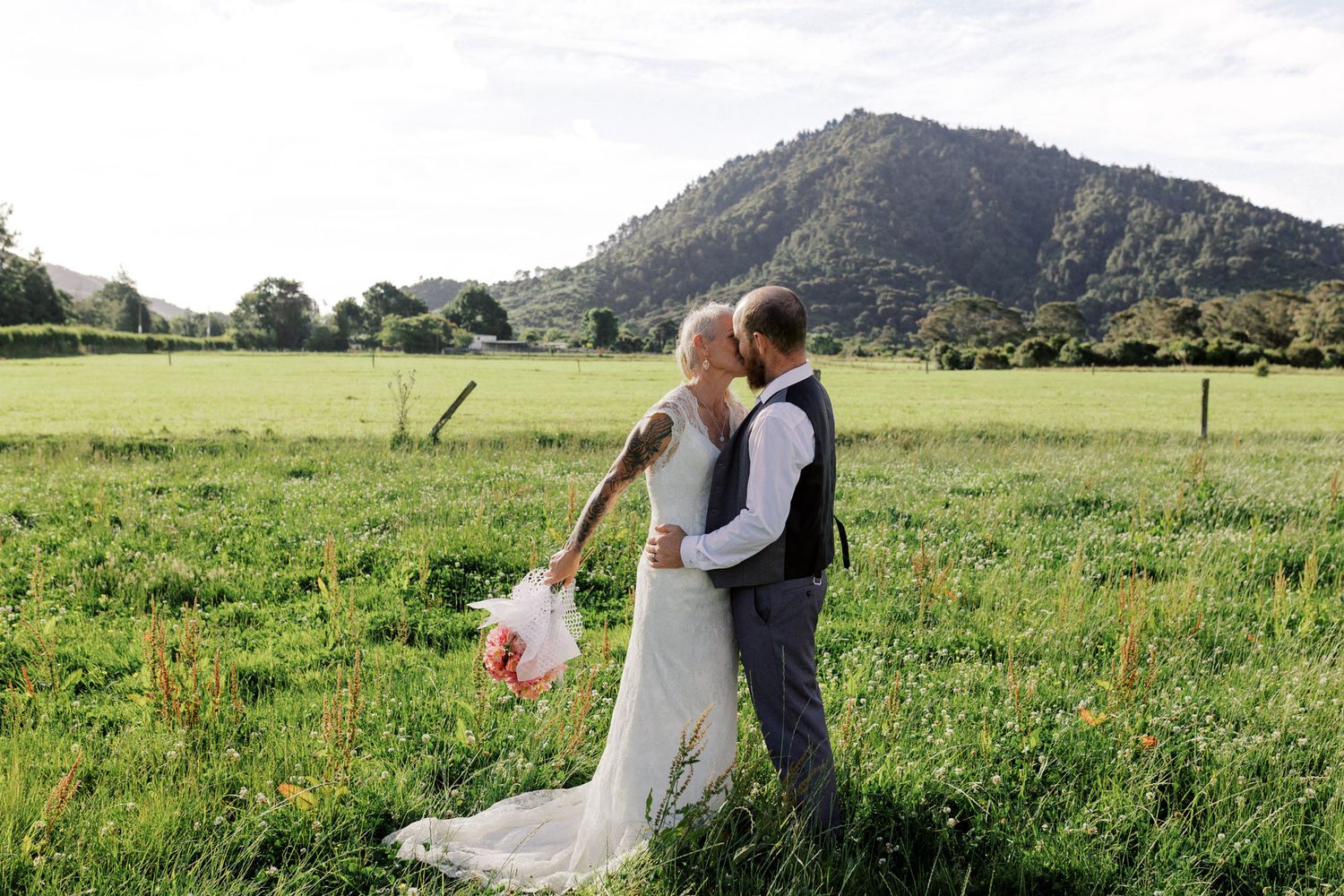 A couple shares a romantic embrace in a lush green meadow with mountains in the background.