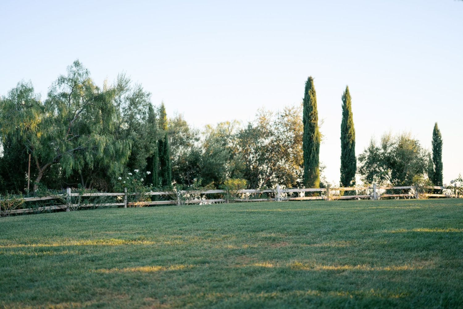 A panoramic view of a rural landscape with cypress trees and white fencing stretches across the countryside.