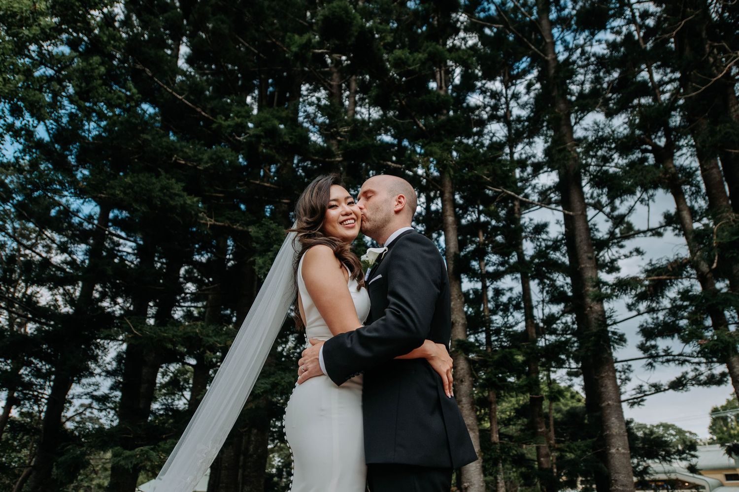 A couple shares a romantic outdoor moment against a backdrop of tall evergreen trees during their wedding.