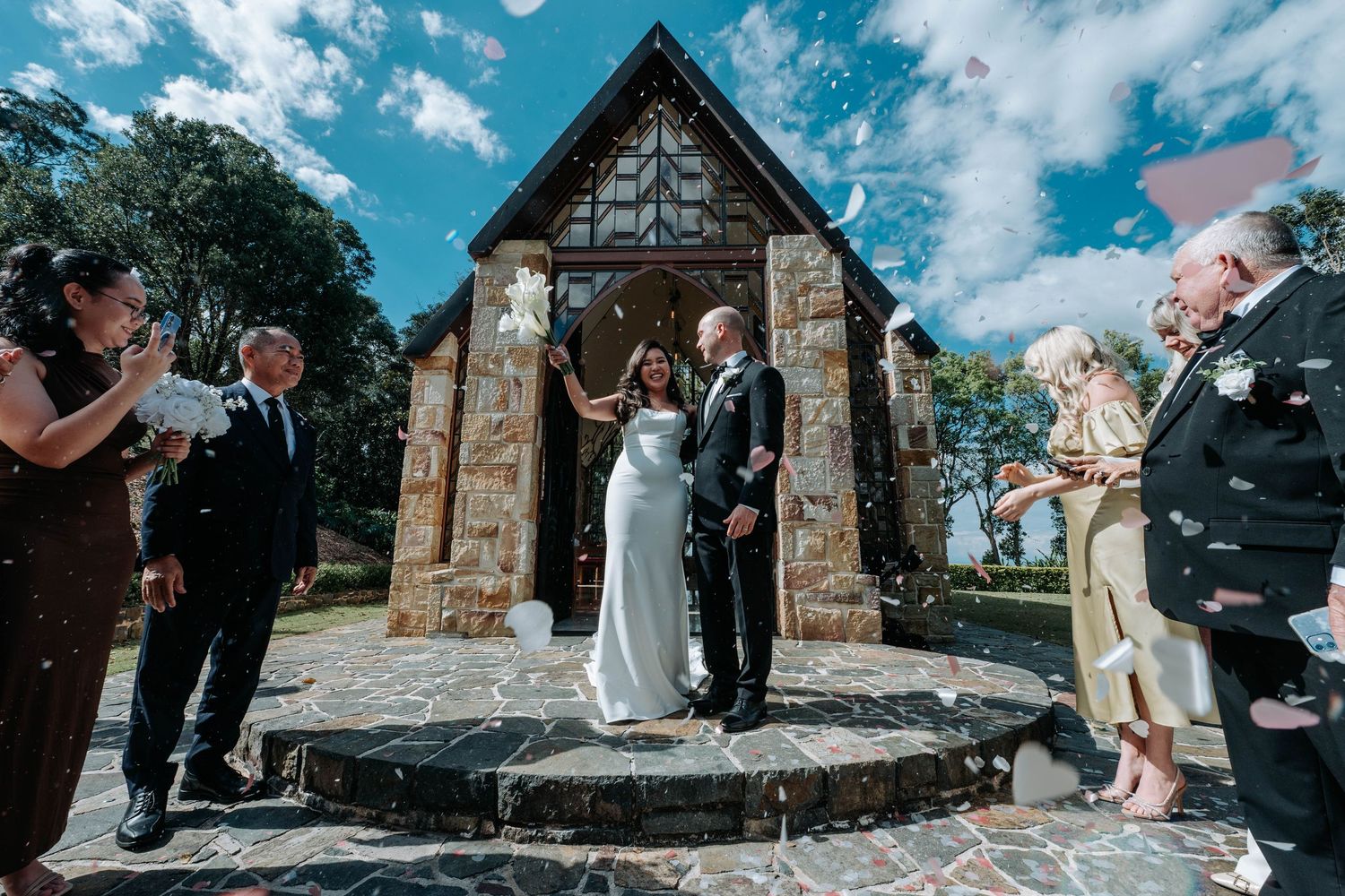 A series of wedding photos taken outside a rustic stone chapel with blue sky and clouds in the background.