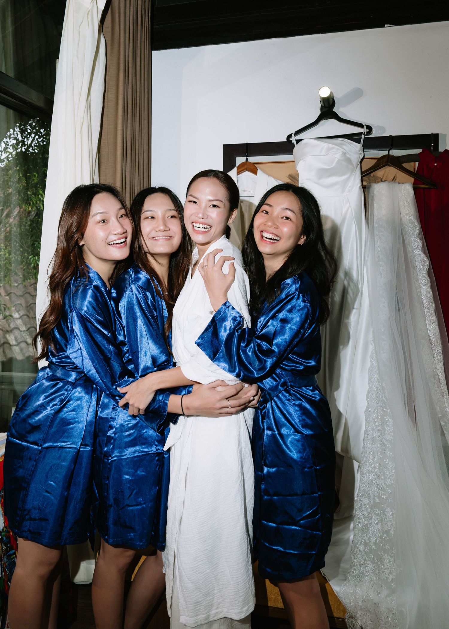 Bride in white robe hugging three bridesmaids in blue satin robes near hanging wedding dress.