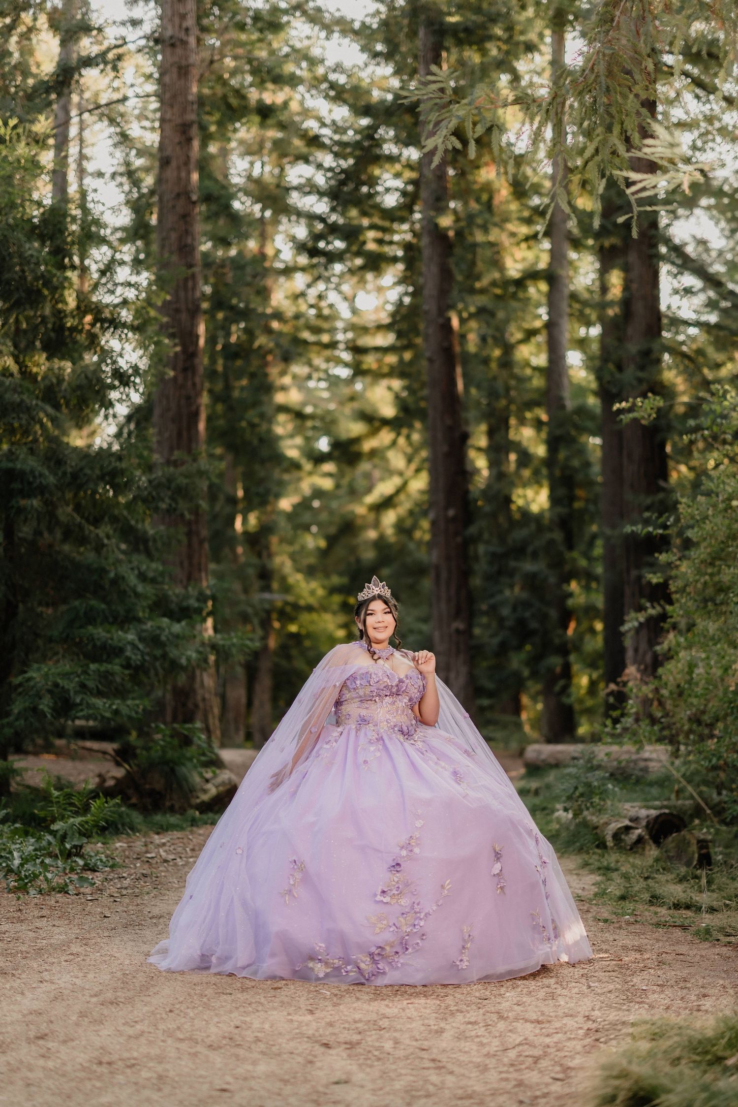A dreamy purple quinceanera dress billows out dramatically on a forest floor surrounded by towering trees.