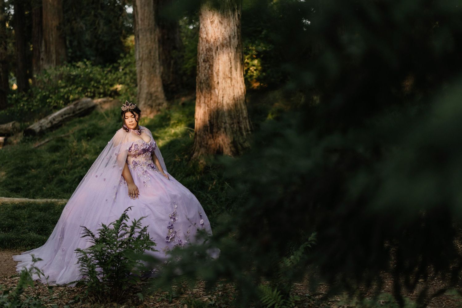 A figure in a lavender ballgown poses gracefully among tall redwood trees in a serene forest setting.