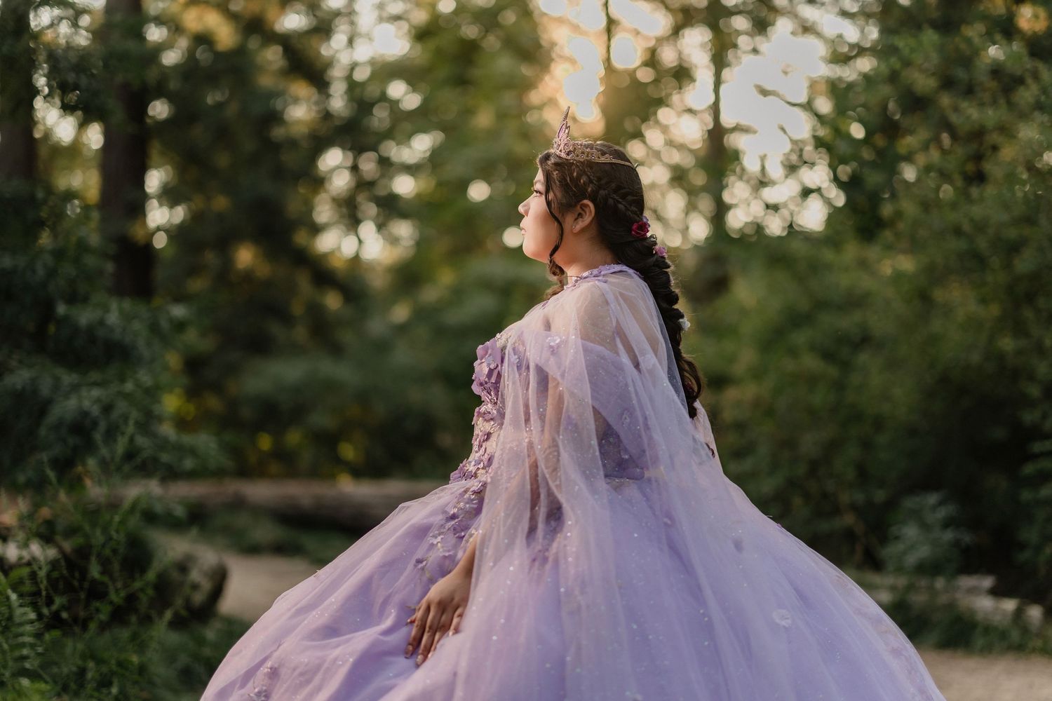 Quinceañera in lavender dress poses outdoors during golden hour in wooded setting.
