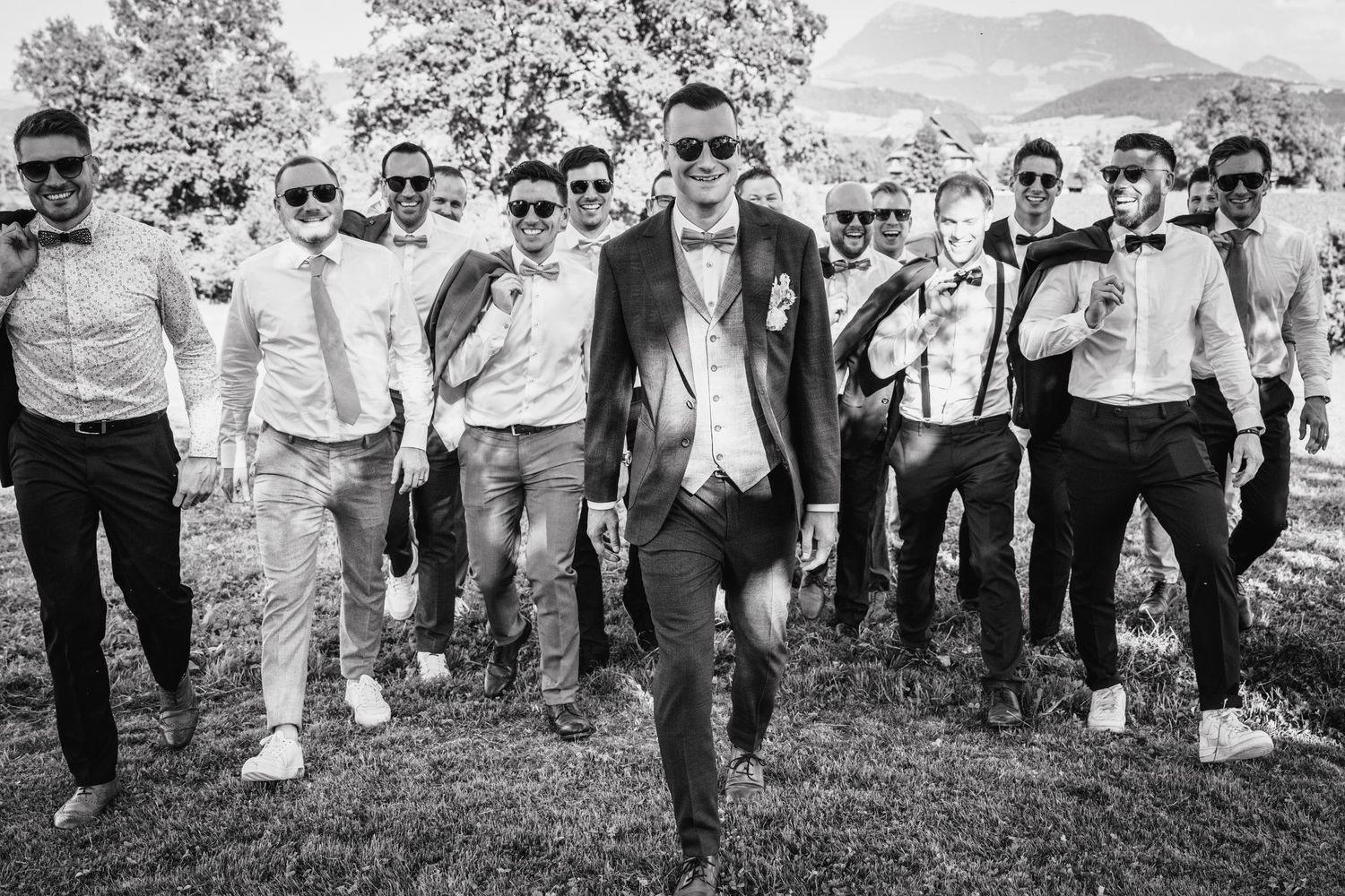 A group of groomsmen stand together on a hillside during a wedding celebration, casting identical poses in black and white.