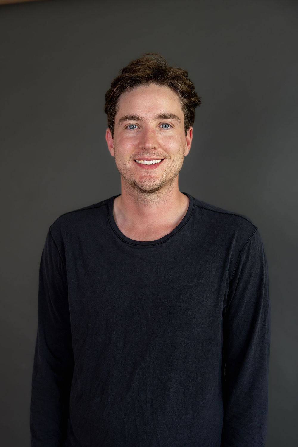 A smiling young man in a dark long-sleeve shirt posing for a casual headshot portrait.