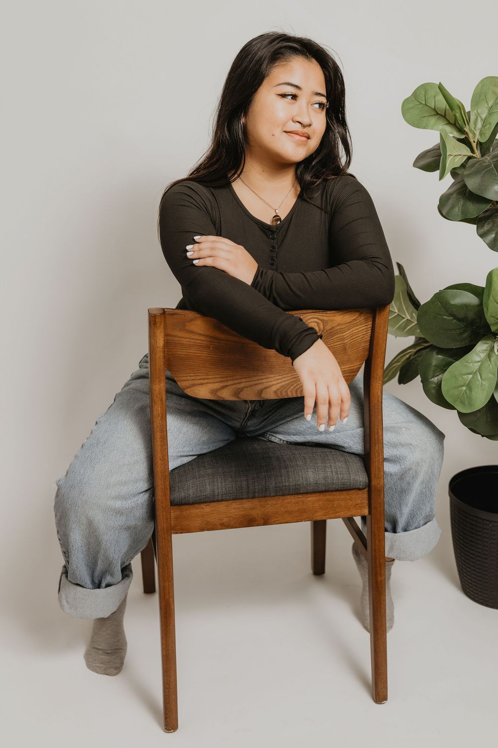 Young woman in dark top and jeans sitting relaxed on wooden chair beside fiddle leaf fig plant.