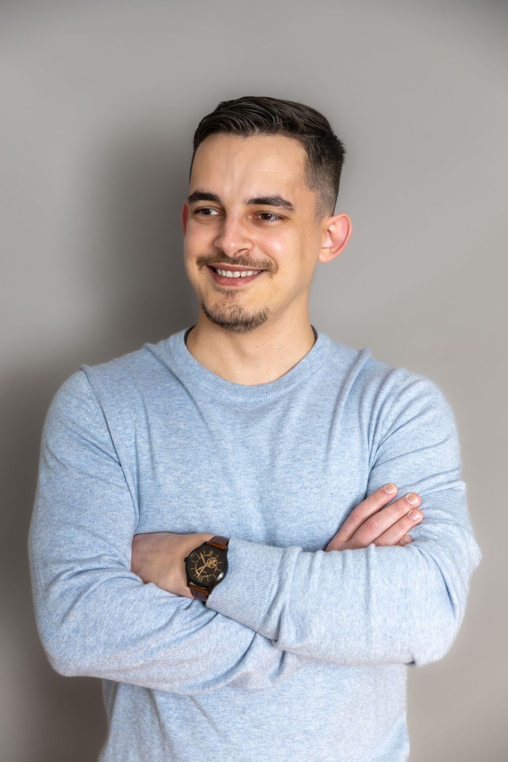 Smiling young man with short dark hair in grey long-sleeve shirt standing with arms crossed.