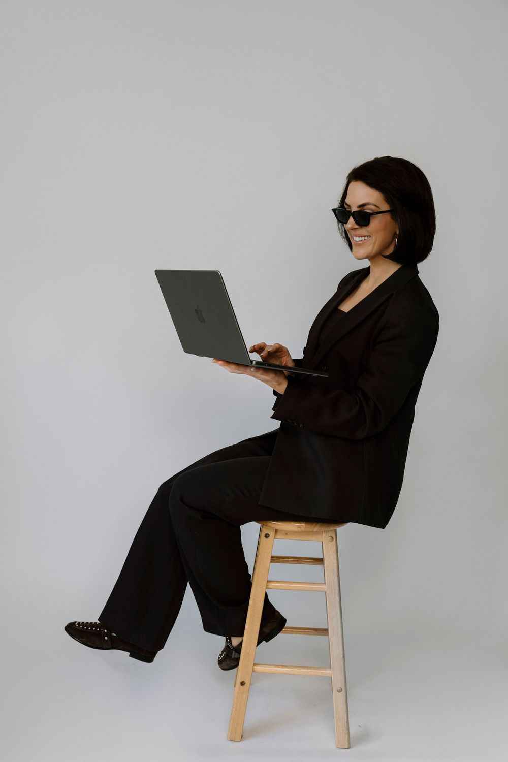 Woman in black suit and sunglasses sitting on wooden stool using laptop, smiling against white background.