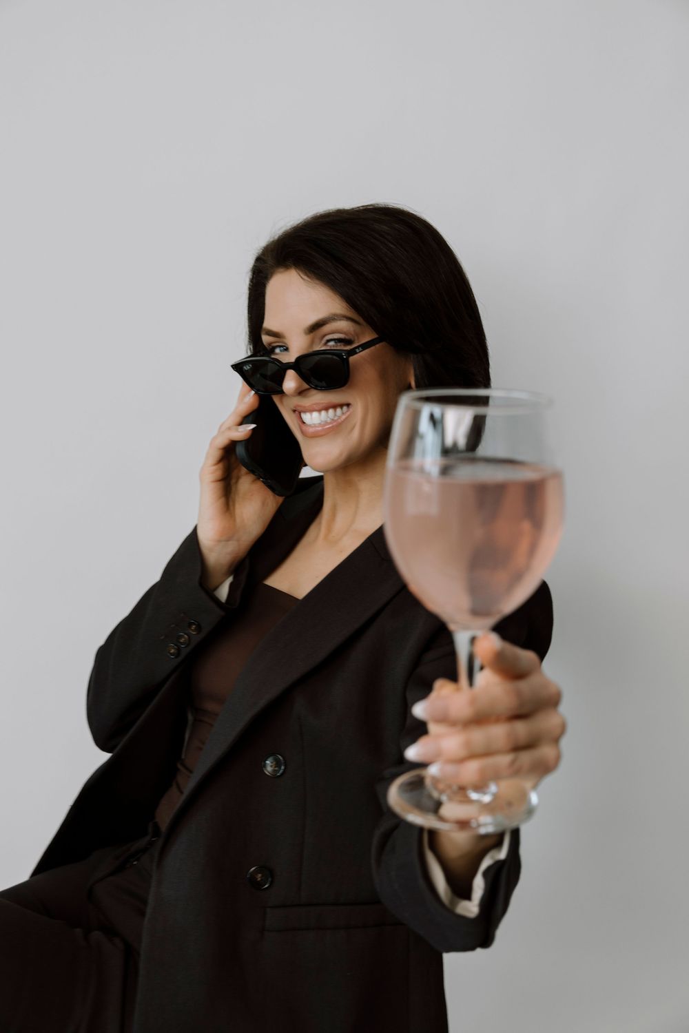 Smiling brunette woman in black blazer and sunglasses holding wine glass while talking on smartphone.