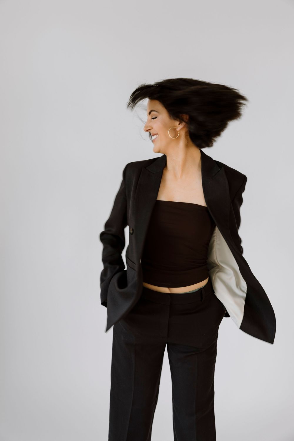 Woman in black blazer and crop top tossing hair, smiling against white background.