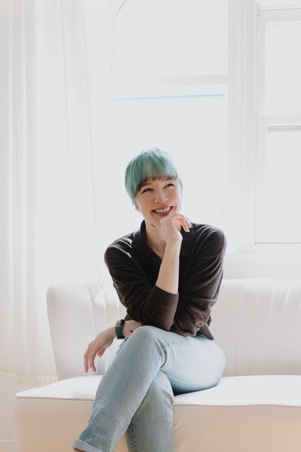 A woman with teal hair sitting on a white couch, smiling warmly in a bright indoor portrait.