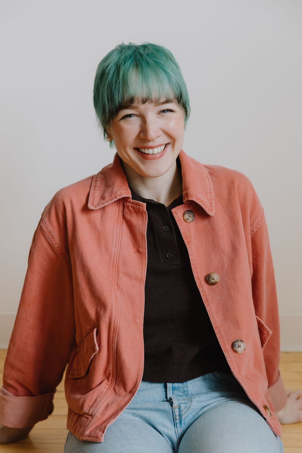 A woman with teal hair wearing a coral jacket smiling in a casual indoor portrait.