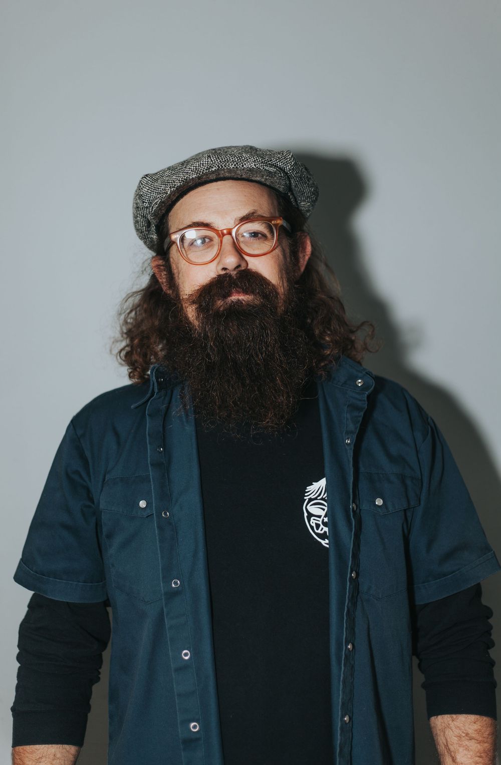 Bearded man wearing glasses and grey flat cap in navy jacket standing against grey background.
