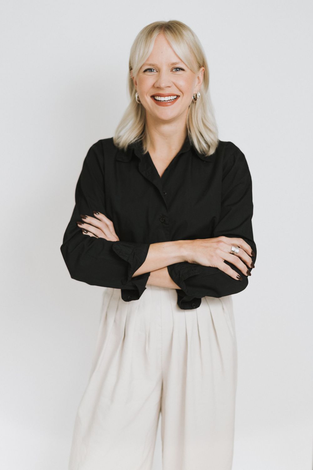 Smiling blonde woman in black shirt and cream trousers standing with arms crossed against white background.