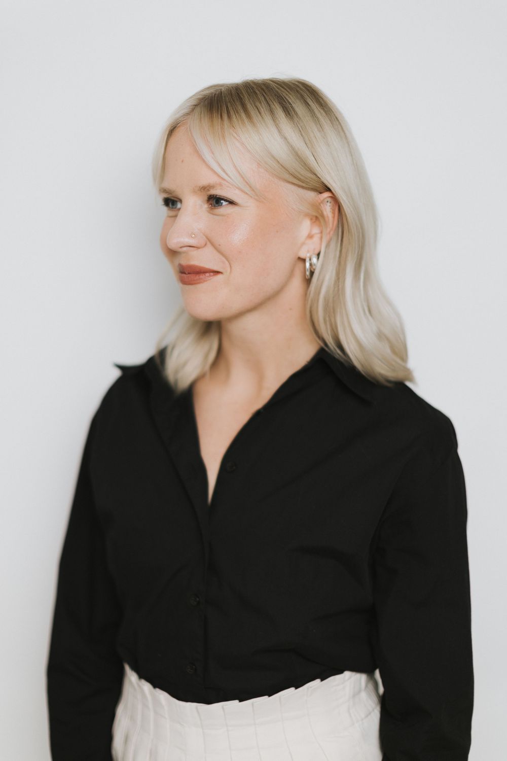 Blonde woman in black button-up shirt looking sideways, wearing hoop earrings against white background.