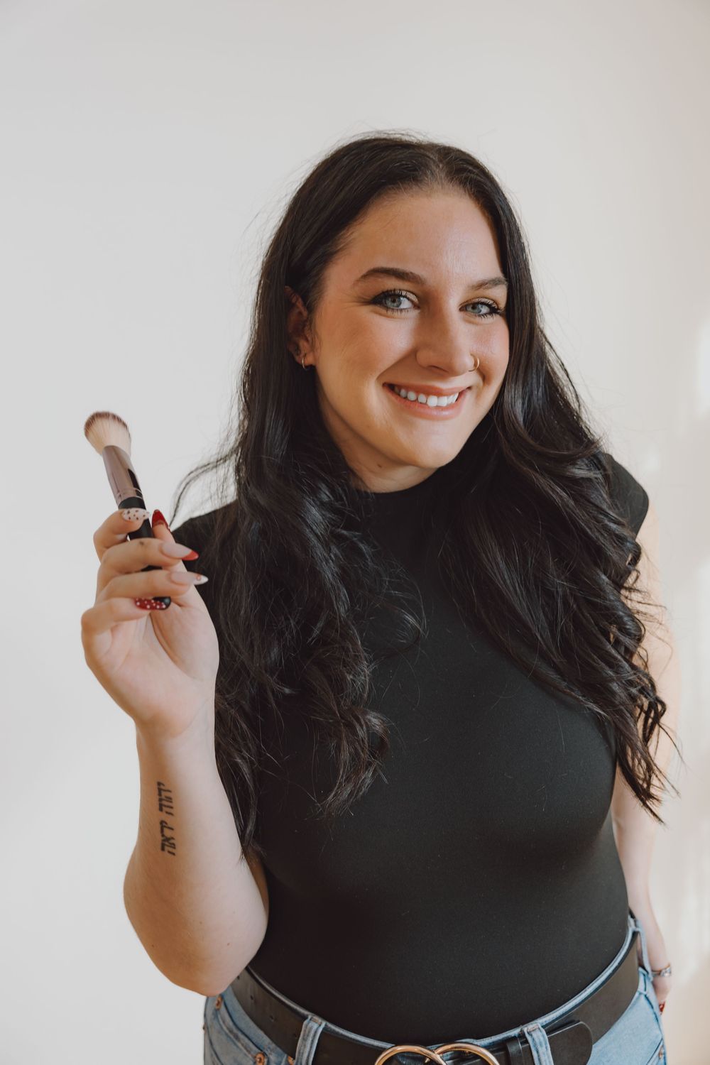 Smiling brunette woman holds makeup brush in black top against white background.