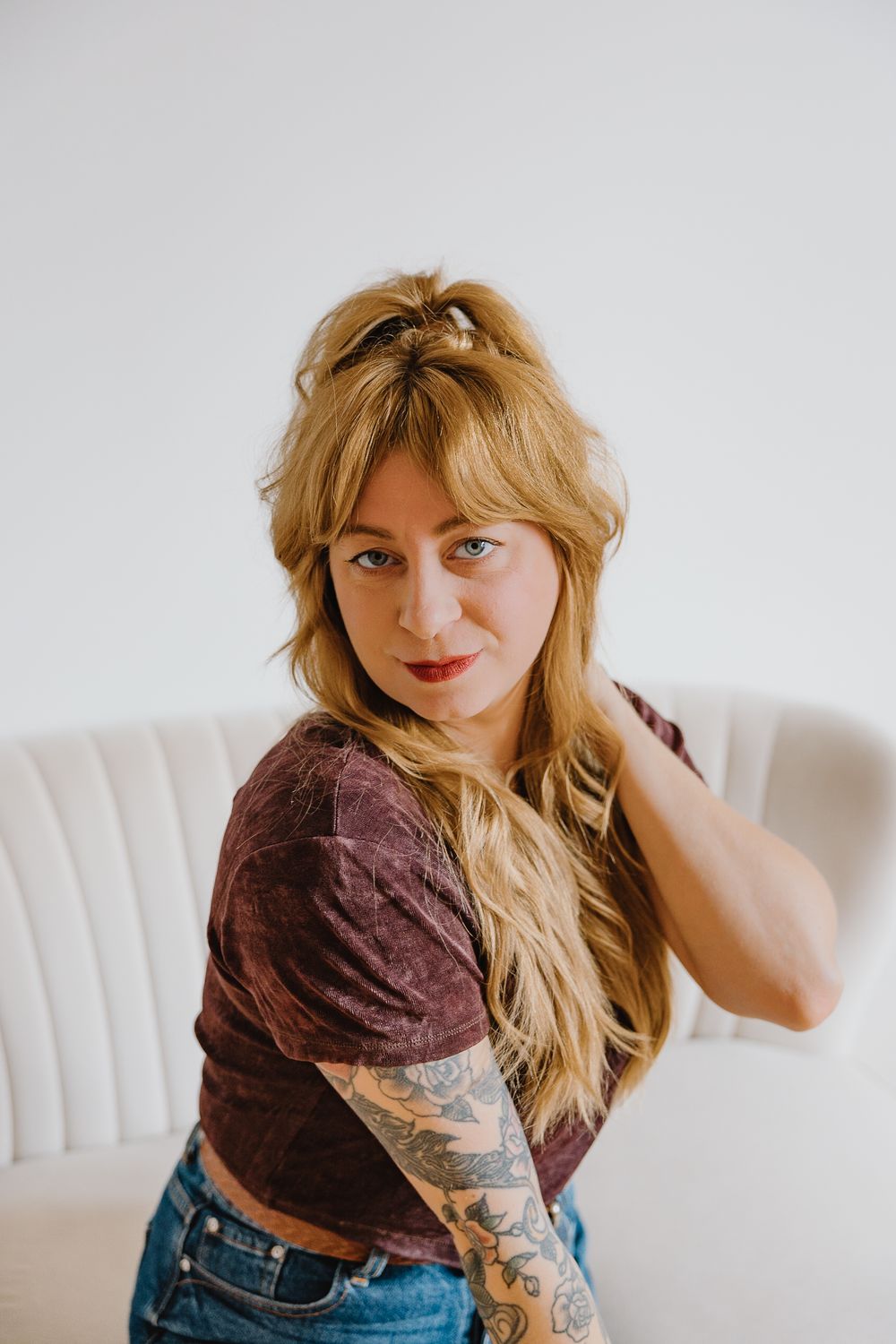 Tattooed blonde woman in burgundy crop top poses on white sofa looking at camera.