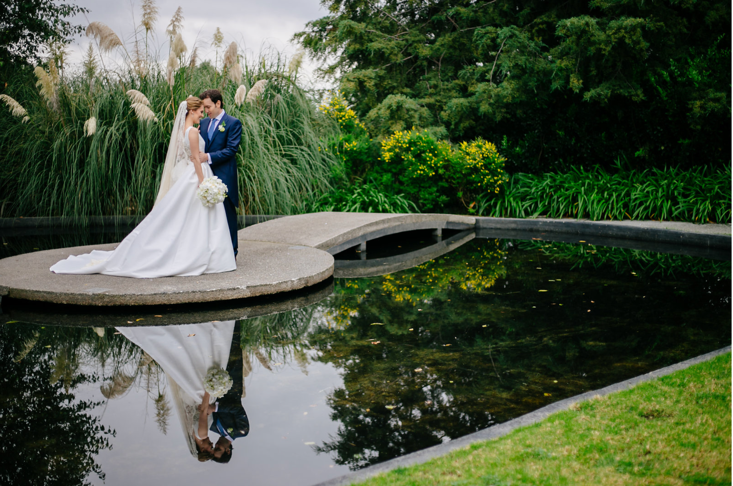 Wedding couple reflected in serene pond surrounded by lush garden landscaping.