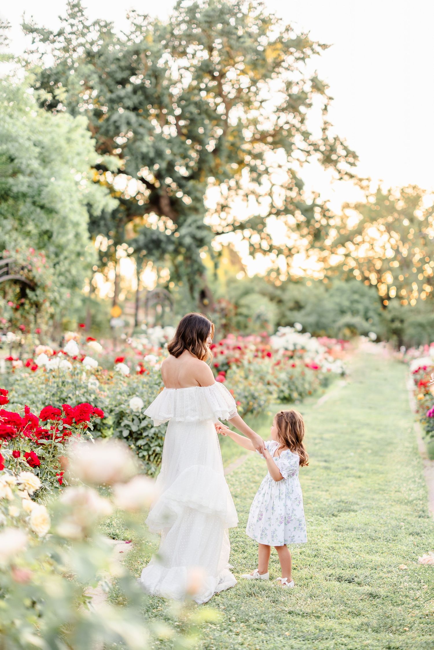 Two people in white dresses dancing through a garden filled with red roses at sunset.