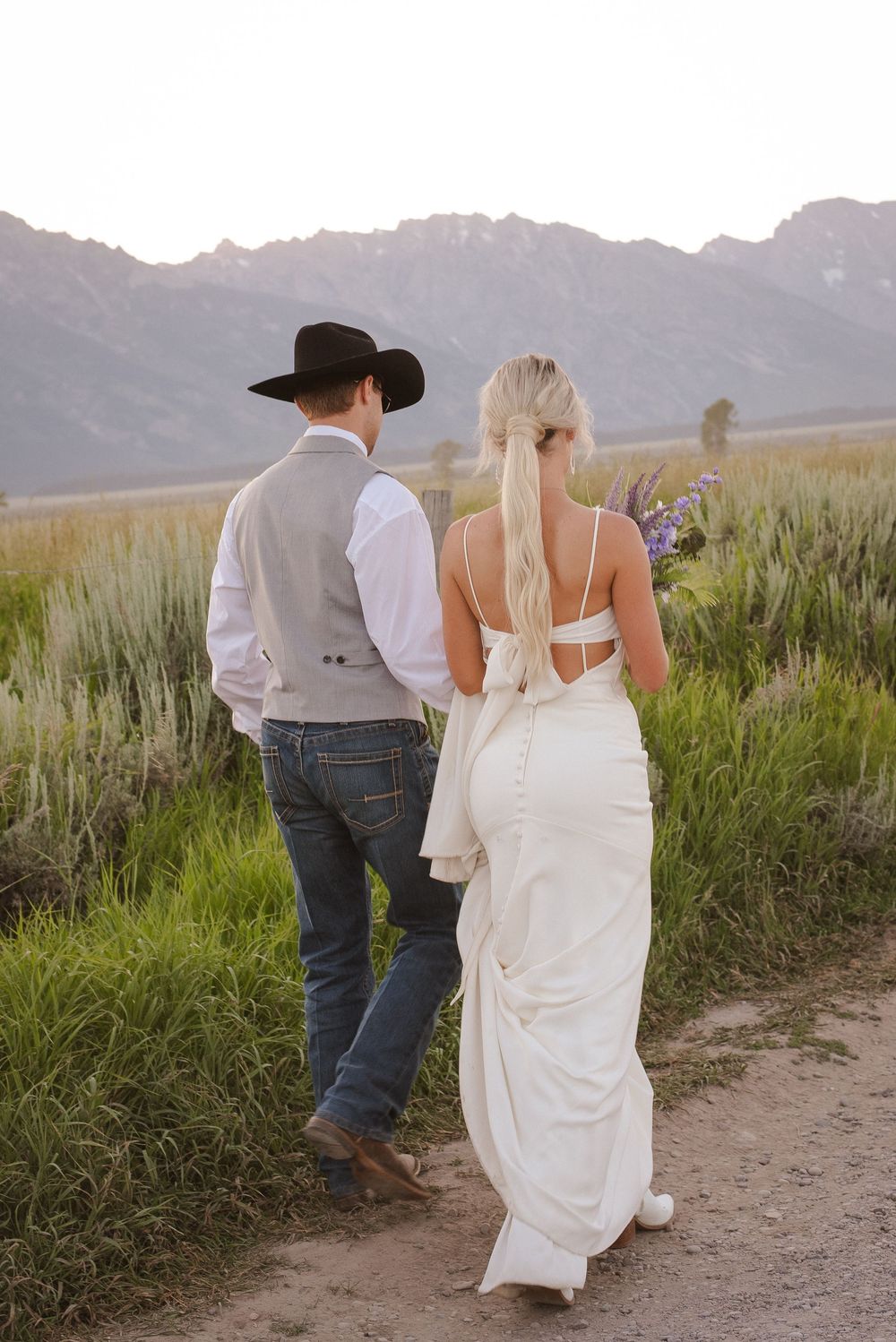 Couple walking through mountain meadow at sunset in western wedding attire.