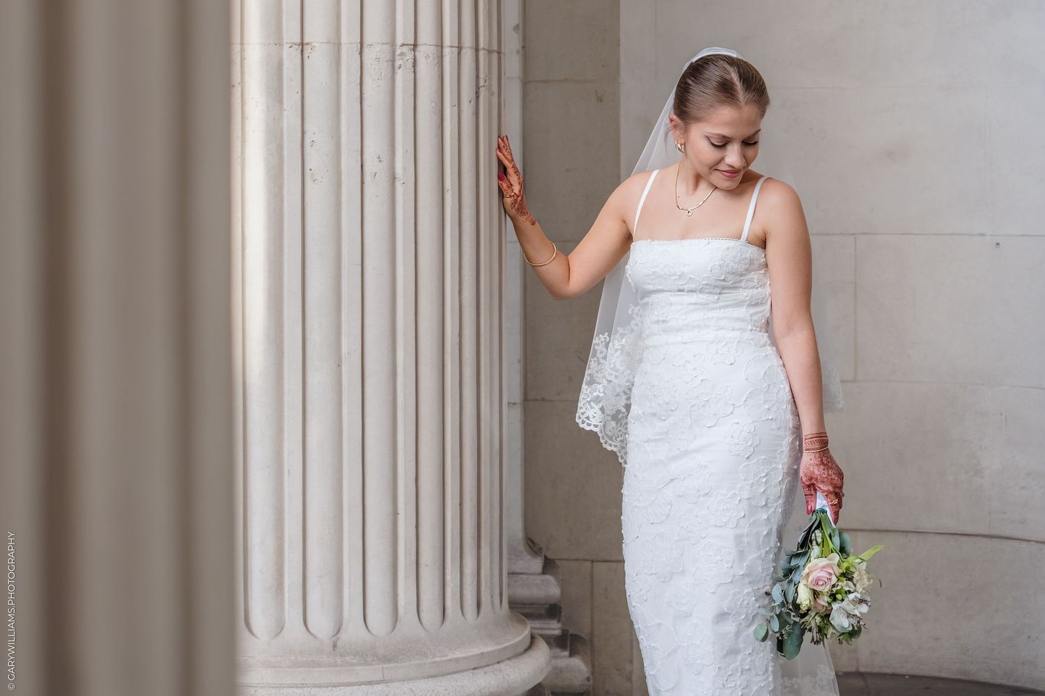 A bride wearing a strapless white wedding gown walks through an elegant columned corridor while carrying a bouquet.