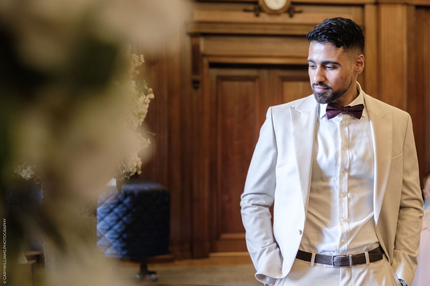 A person in a light-colored suit stands in a warmly lit wooden-paneled room with elegant decor.