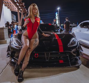 Tattooed model in a red bodysuit sitting on the hood of a black sports car with red racing stripes at a nighttime car meet.