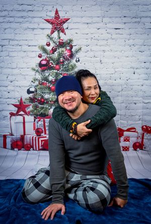 Smiling couple in festive pajamas posing with a piggyback in front of a decorated Christmas tree for a holiday photoshoot.