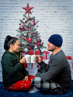 Couple in cozy winter pajamas sitting by a decorated Christmas tree sharing hot drinks during a holiday photo session.