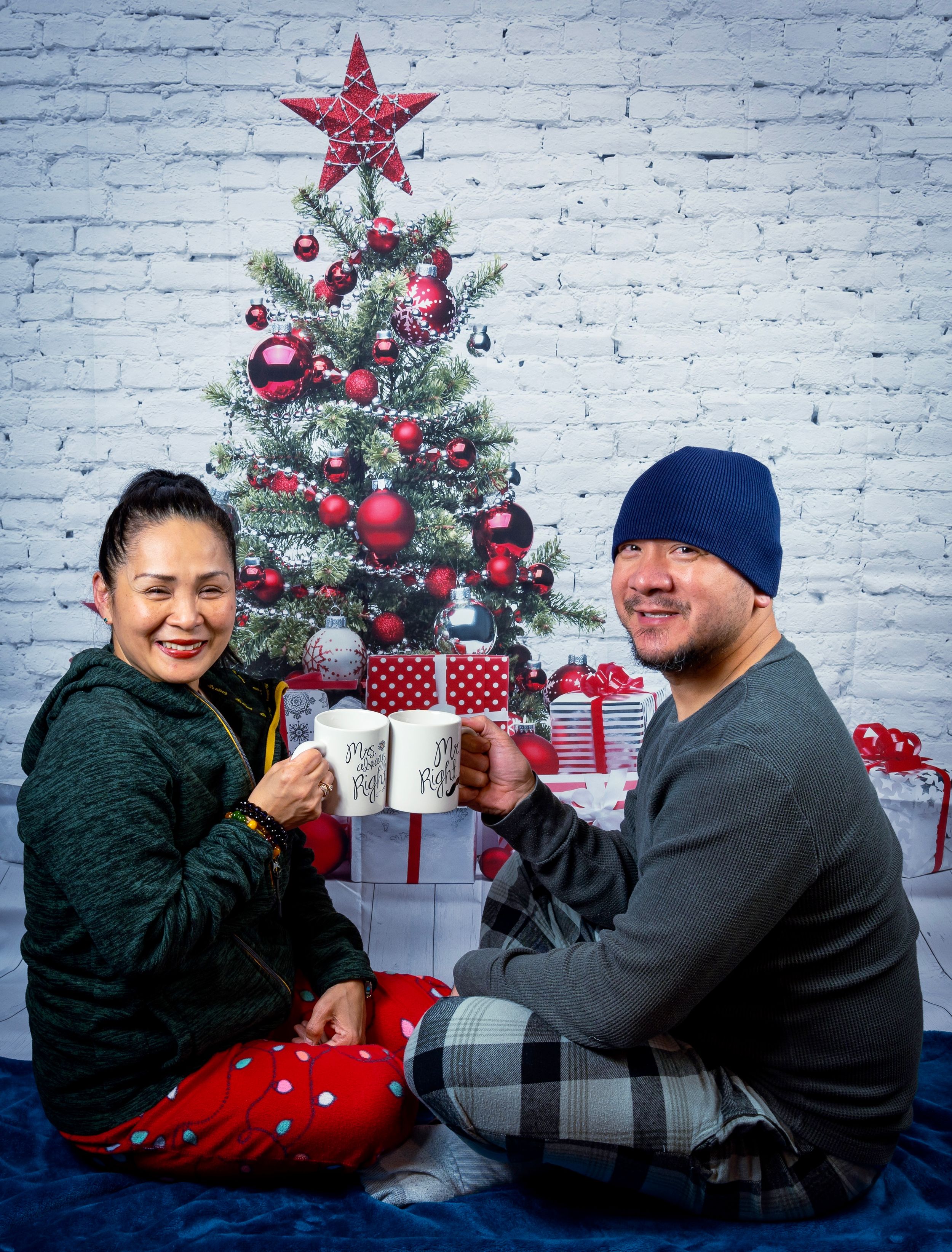 Couple in festive pajamas sitting by a decorated Christmas tree holding mugs during a cozy holiday photoshoot.