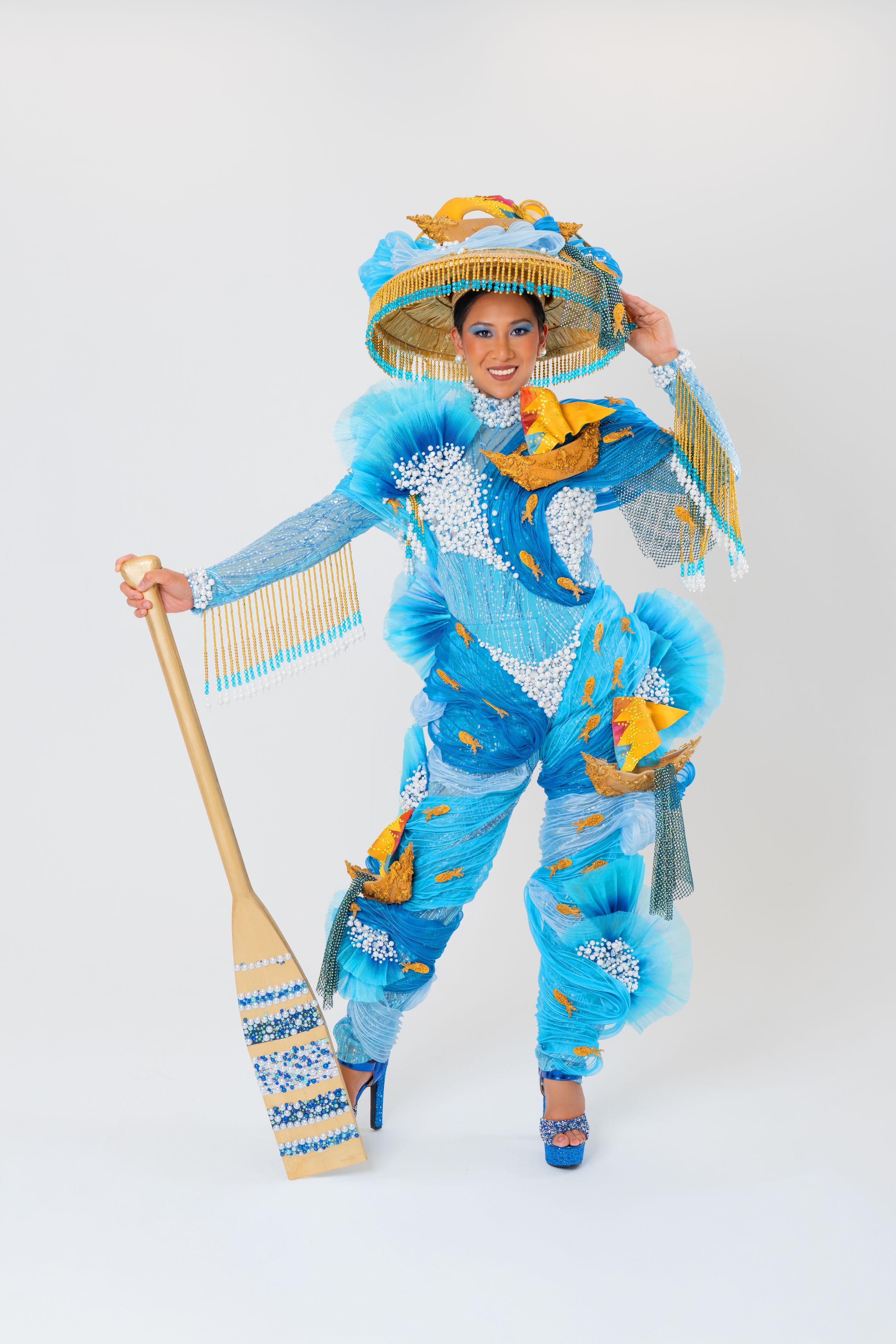 Full-body view of a model posing in a blue and gold beaded costume with a matching hat and decorative paddle in a white studio.