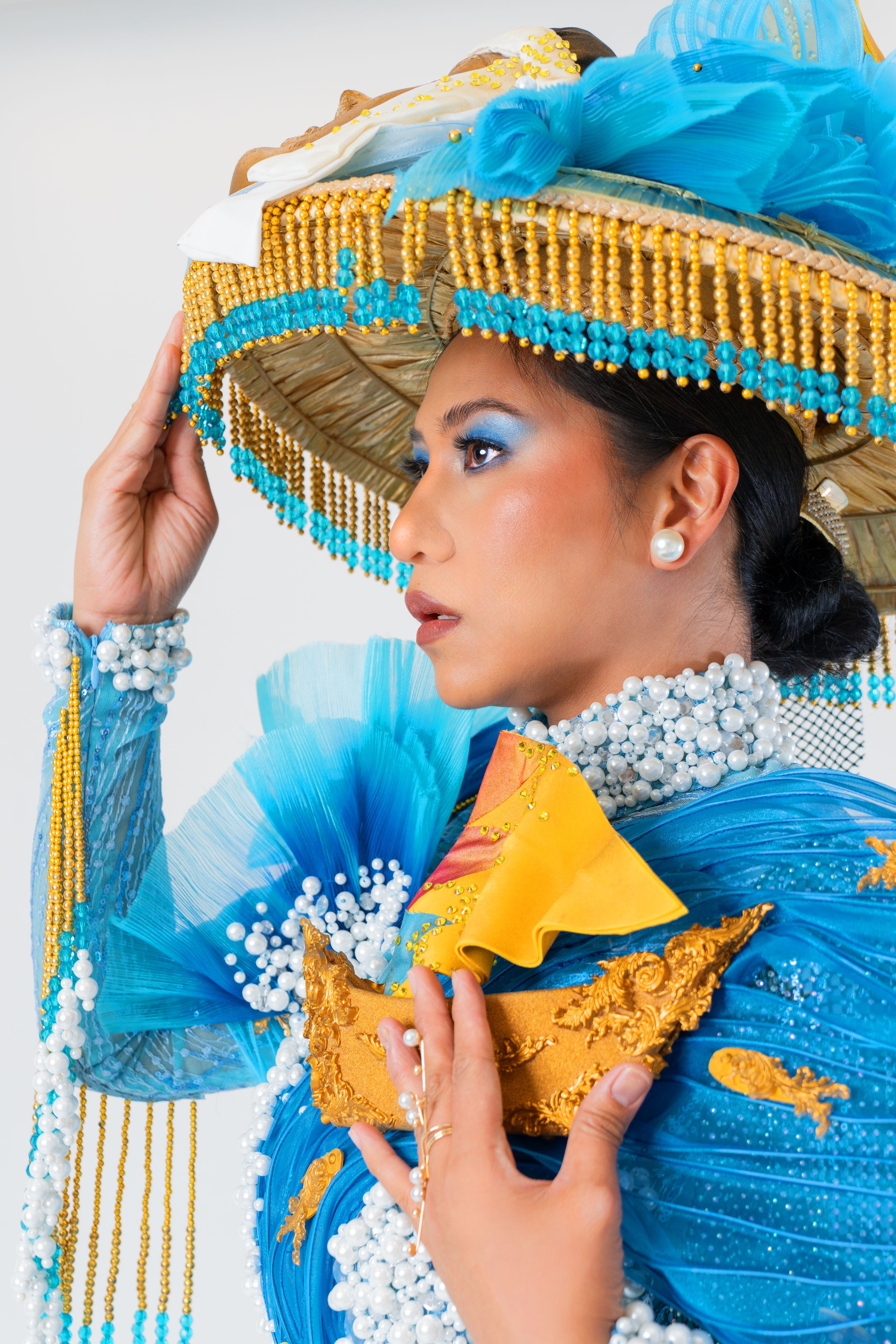 Side portrait of a model in a blue and gold culturally-inspired outfit with matching wide-brim hat accented with beads and water elements.