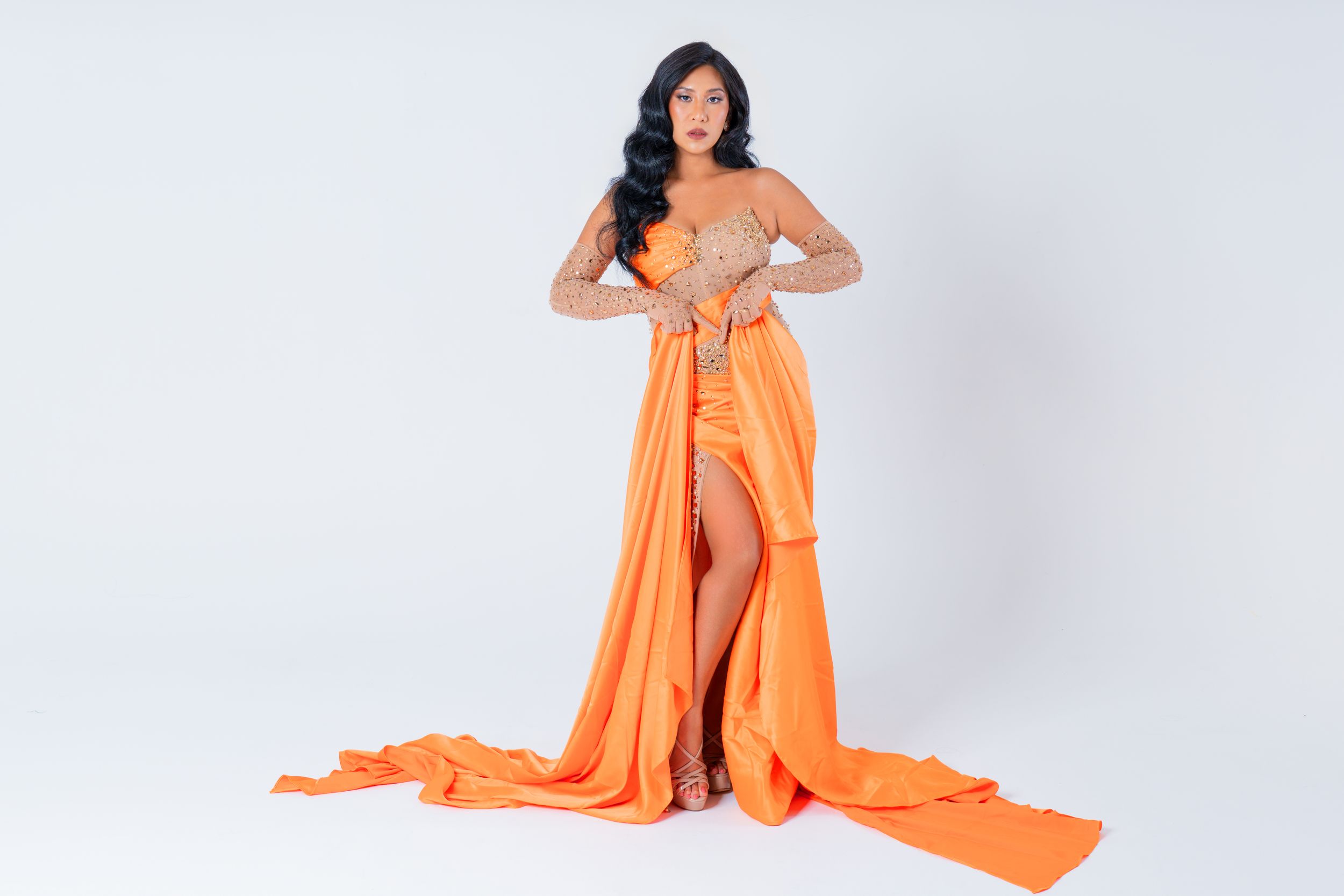 Fashion model posing in a strapless orange evening gown with long flowing train in a white studio.