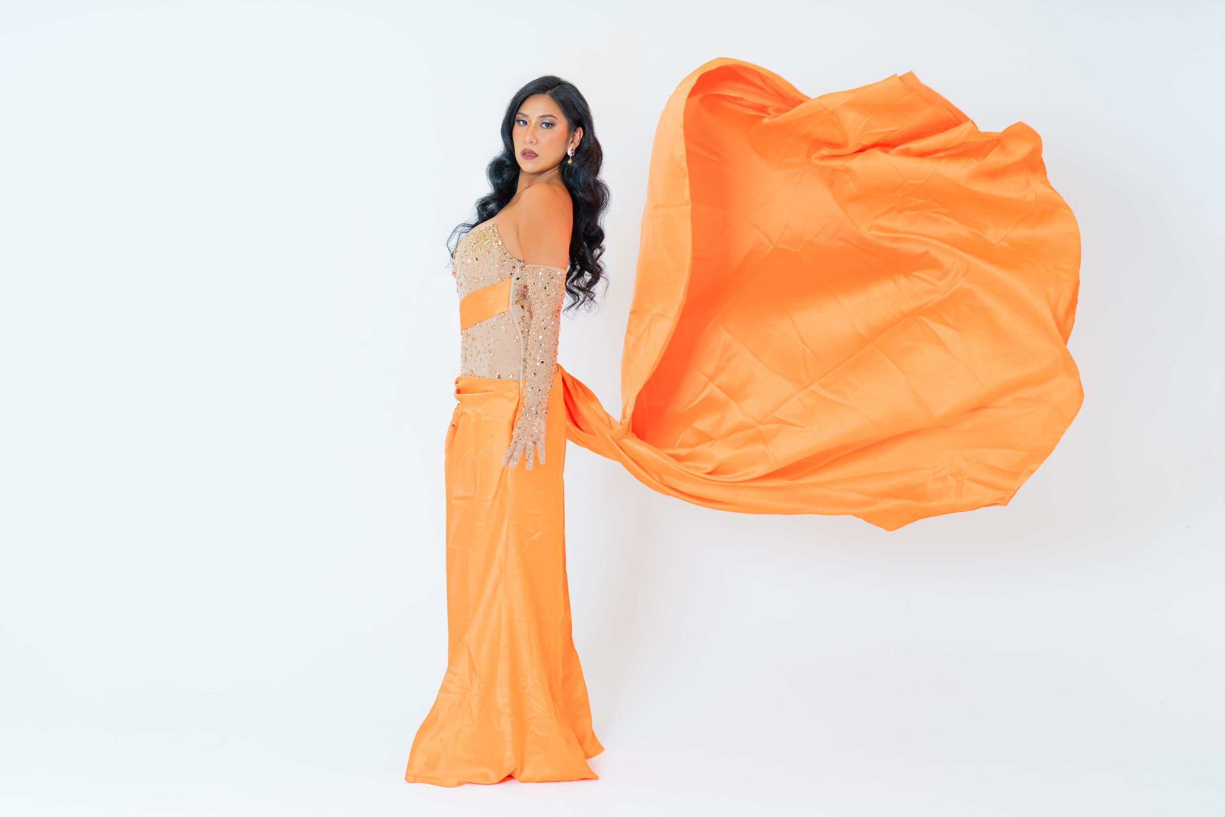 Model in an elegant orange designer gown tossing a dramatic train for a high-fashion studio photoshoot.