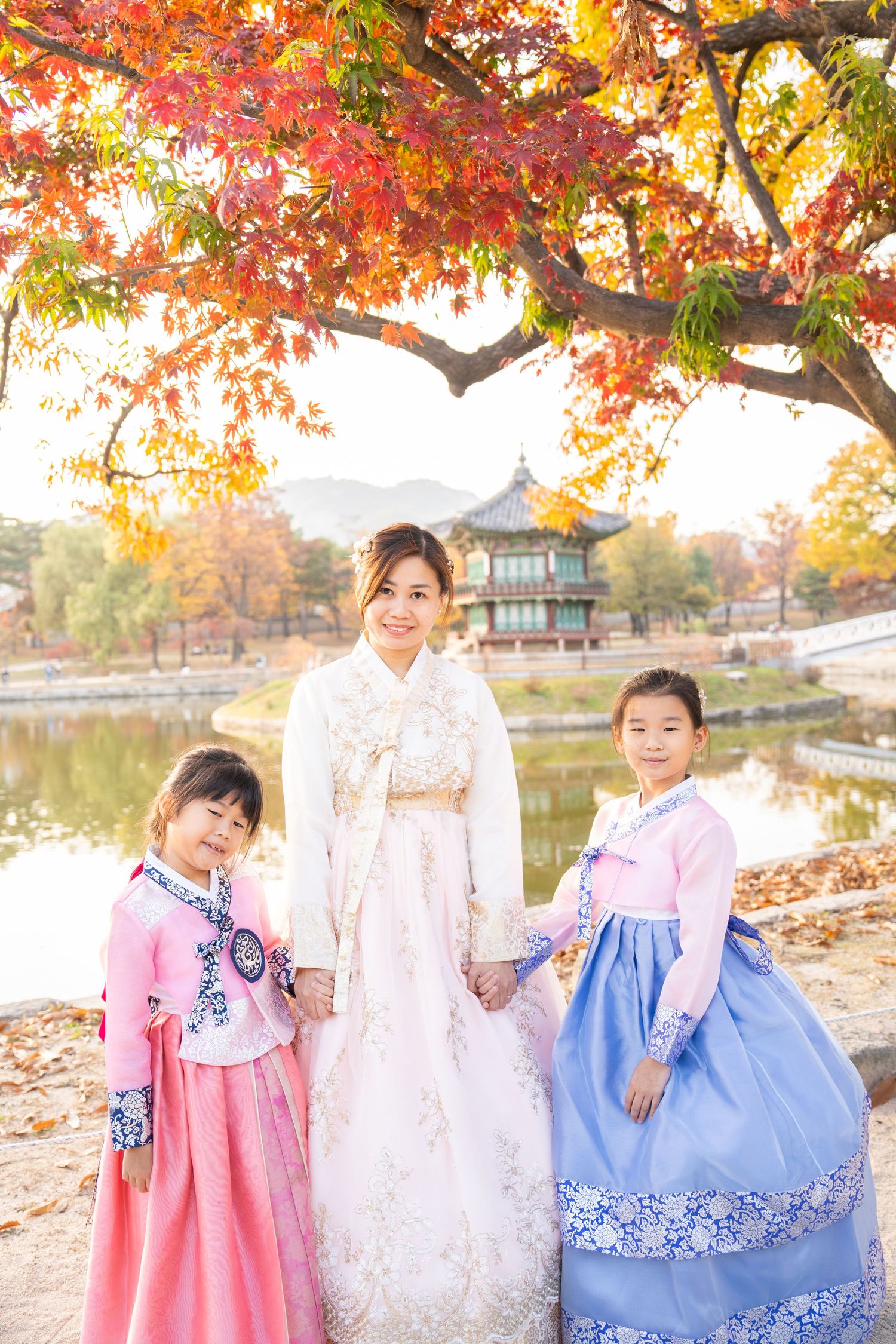 Three people wearing traditional Korean hanbok dresses pose in front of autumn trees and pagoda by a lake.