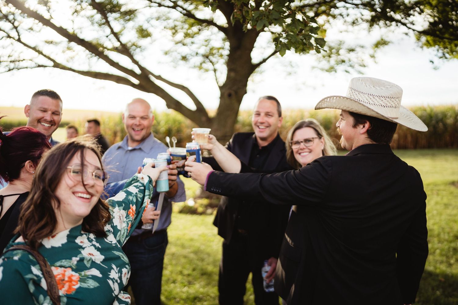 A group of people outdoors enjoying drinks and laughing together in a green field at sunset.