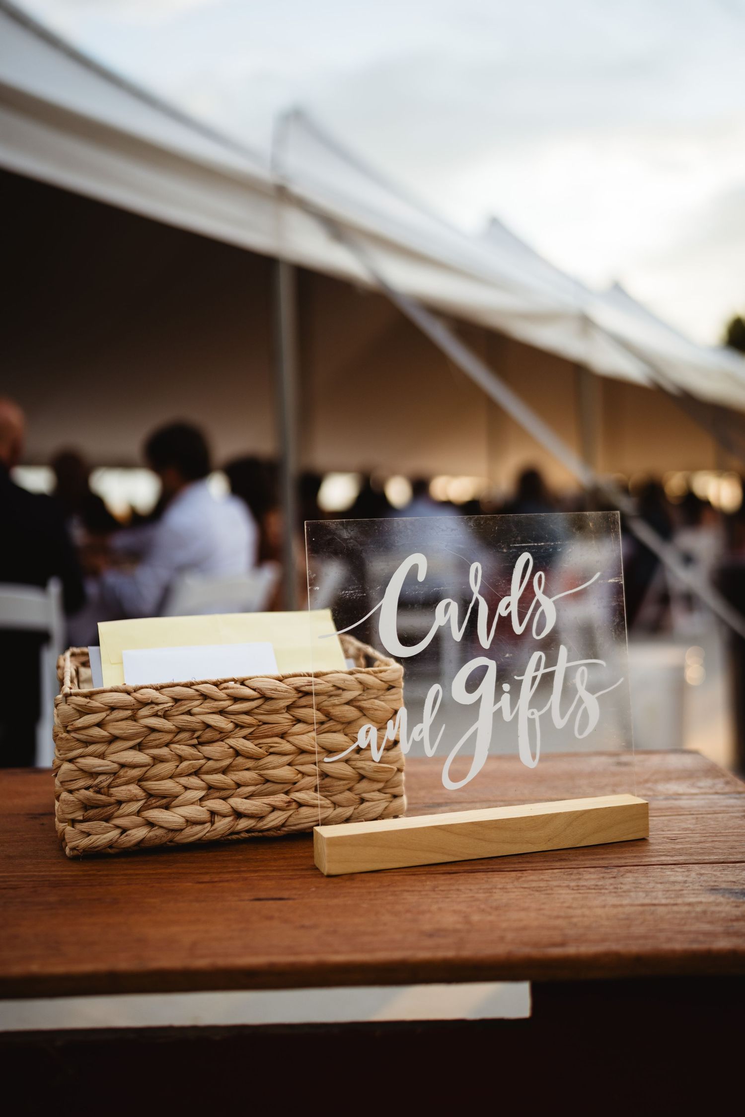 Clear acrylic wedding card signs displayed alongside woven basket card holders on a wooden table at a reception venue.
