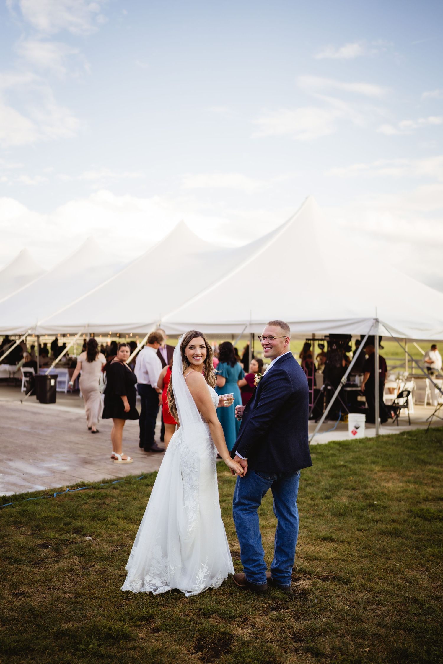 Wedding reception setup outdoors with white tents and guests gathering on the grass at sunset.